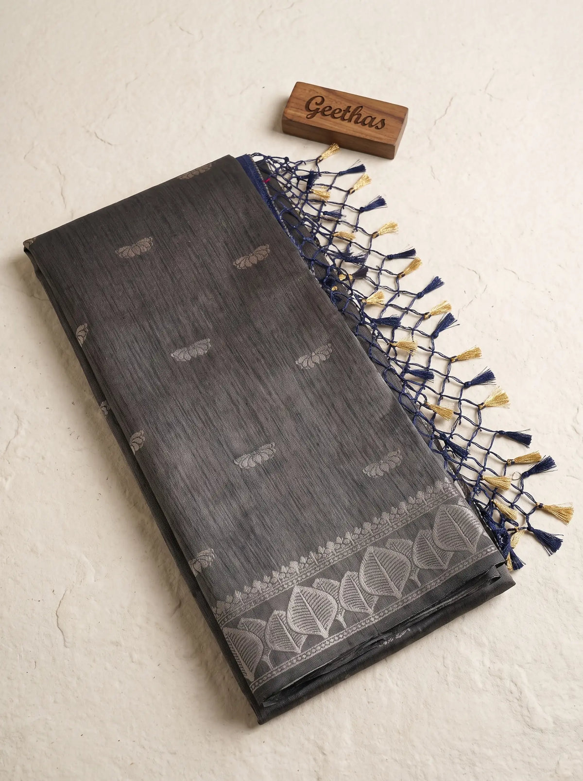 Folded charcoal grey soft tissue saree with woven leaf motifs, leaf border and navy-gold tassled pallu