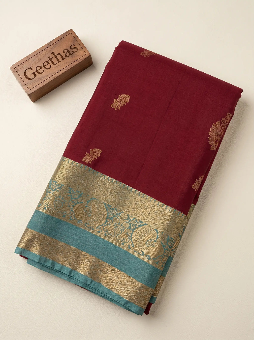 Folded maroon soft silk saree showing gold-toned woven motifs and turquoise blue decorative border