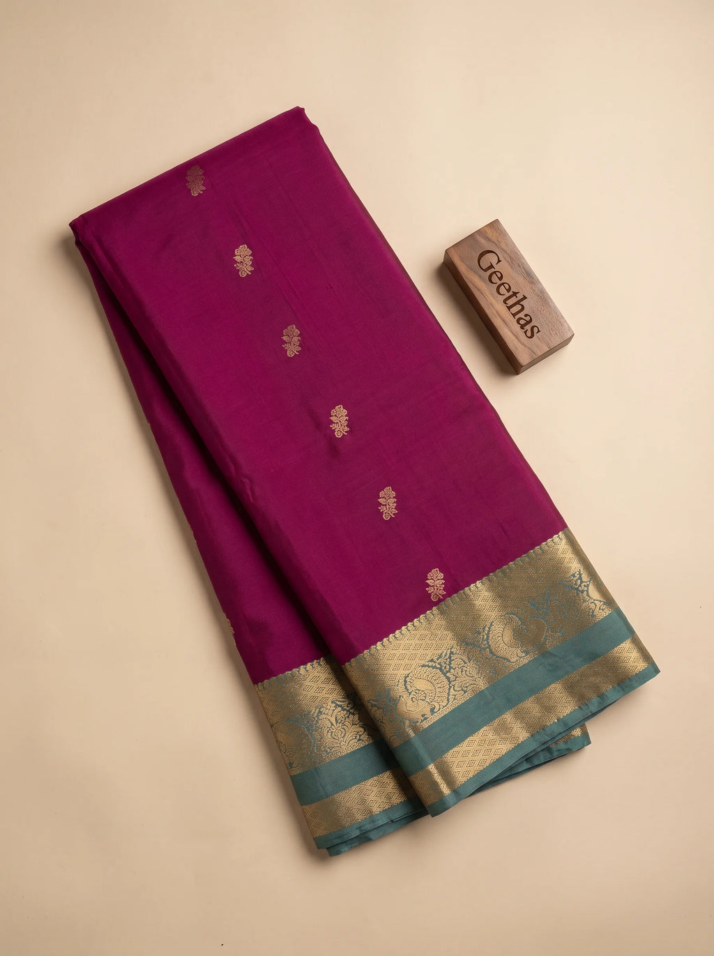 Folded magenta soft silk saree with small gold-toned floral motifs and teal decorative woven border