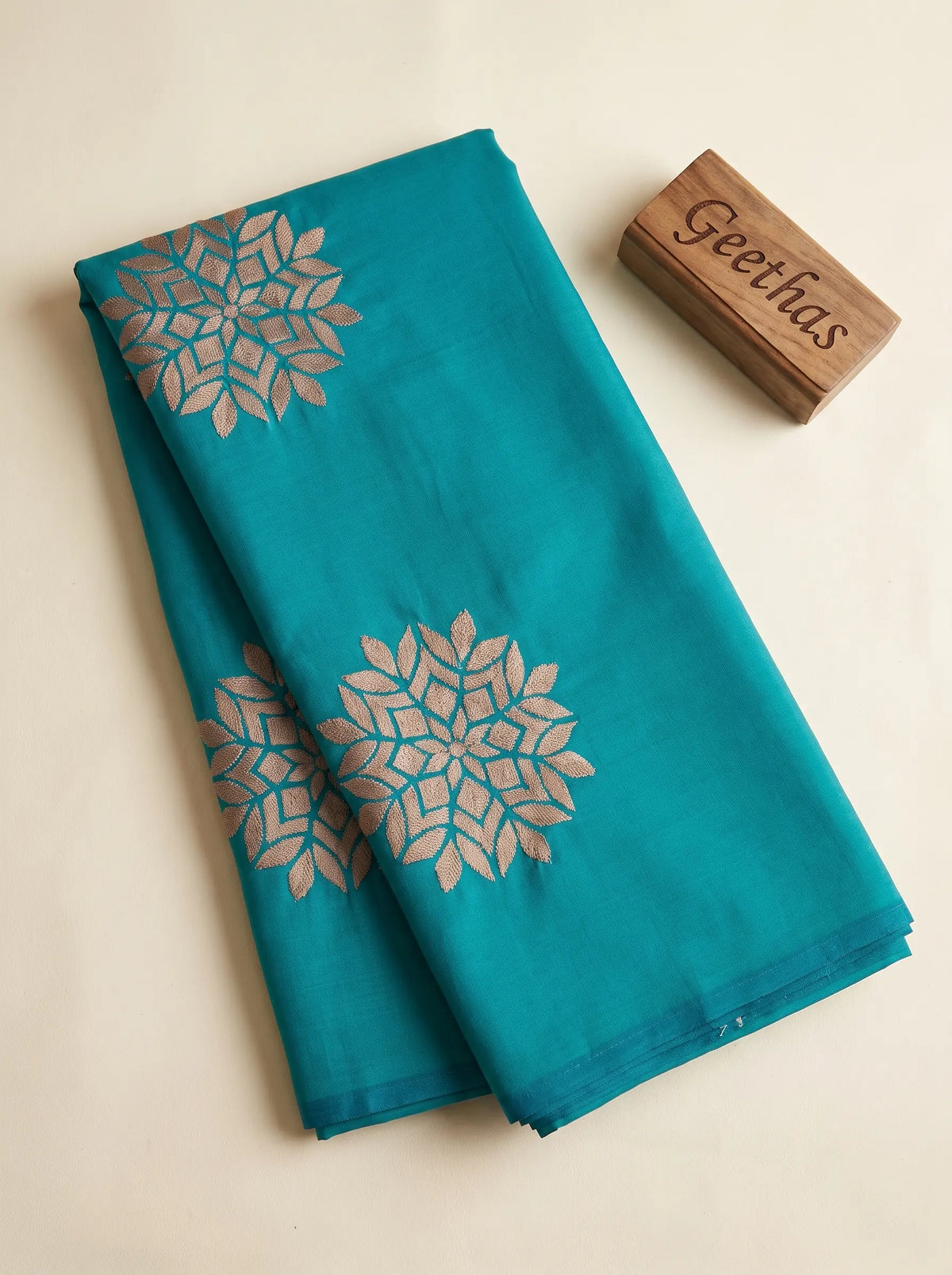 Teal blue soft silk saree folded to display muted gold-toned woven floral-geometric motifs