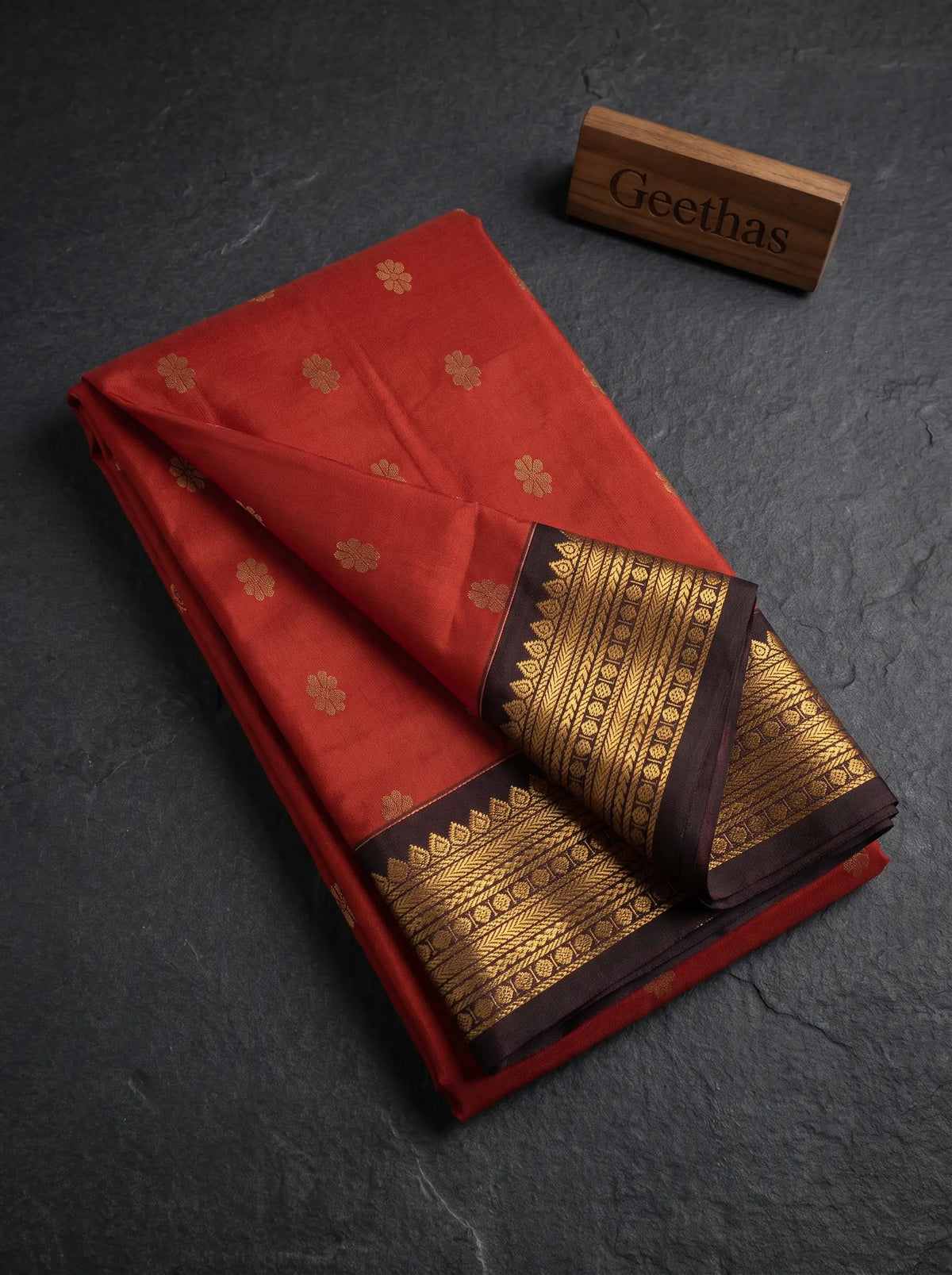 Folded rust orange soft silk saree with gold-toned floral motifs and deep brown zari border