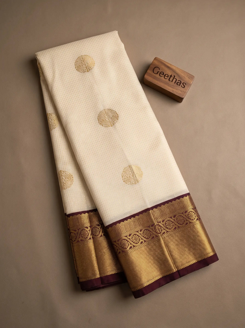 Folded half white soft silk saree with gold-toned circular motifs and a brown decorative border