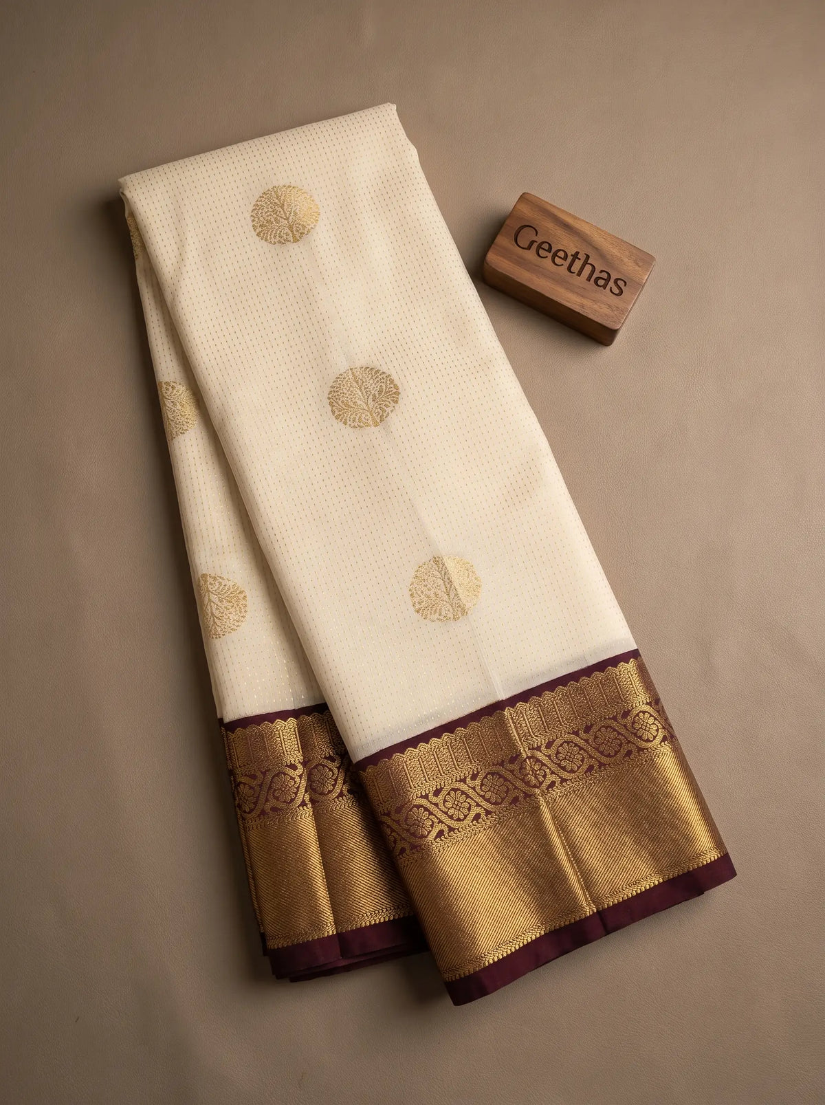 Folded half white soft silk saree with gold-toned circular motifs and a brown decorative border