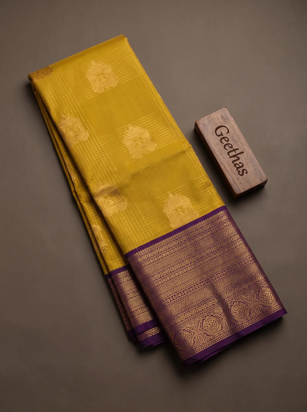 Folded mustard yellow soft silk saree with gold-toned woven motifs and a deep purple zari border.