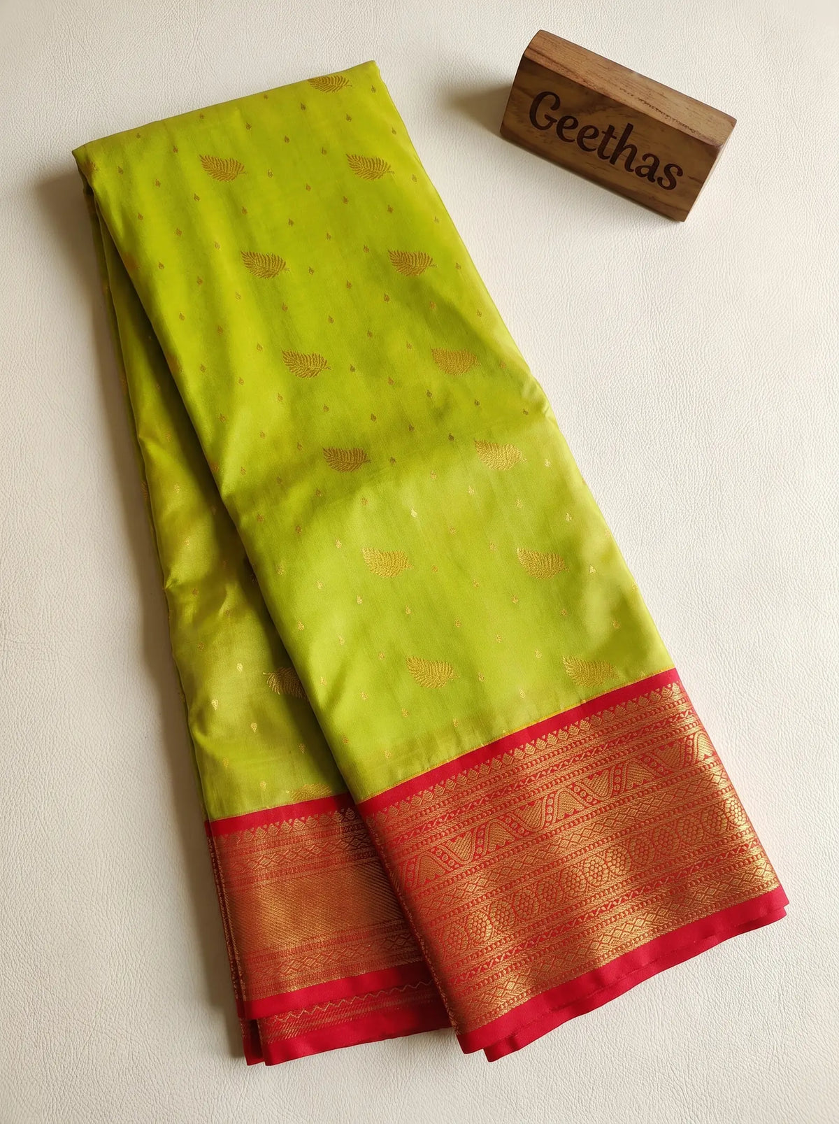 Bright olive soft silk saree with gold leaf motifs and a red gold-patterned border, displayed folded
