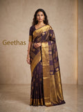 Model wearing a deep brown soft silk saree with gold-toned floral motifs and a broad decorative woven border