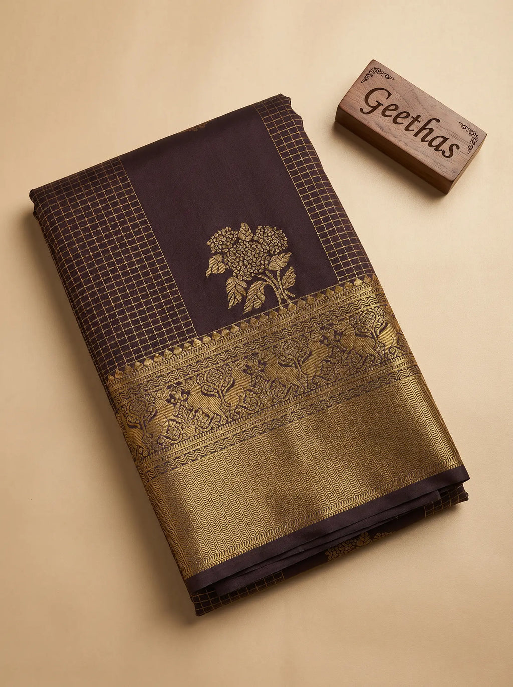 Folded deep brown soft silk saree showing gold-toned floral motifs, geometric check panels and detailed woven border