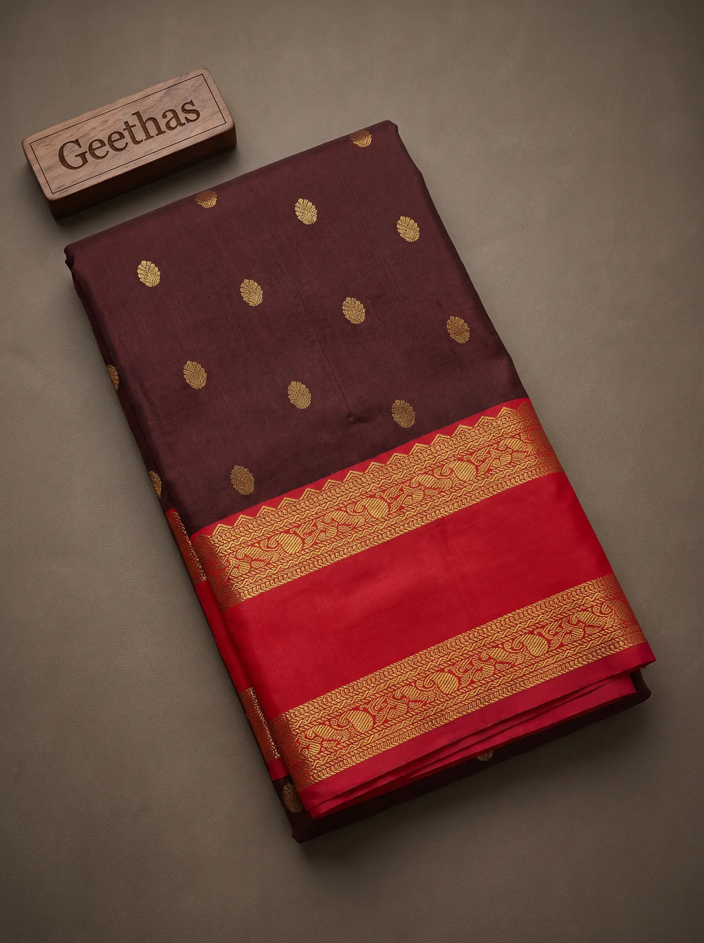 Folded deep brown soft silk saree with gold-toned woven motifs and a red decorative zari-style border
